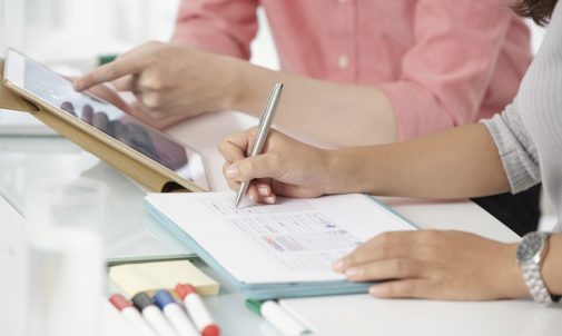 Faceless shot of contemporary colleagues using tablet while writing on clipboard sitting at table